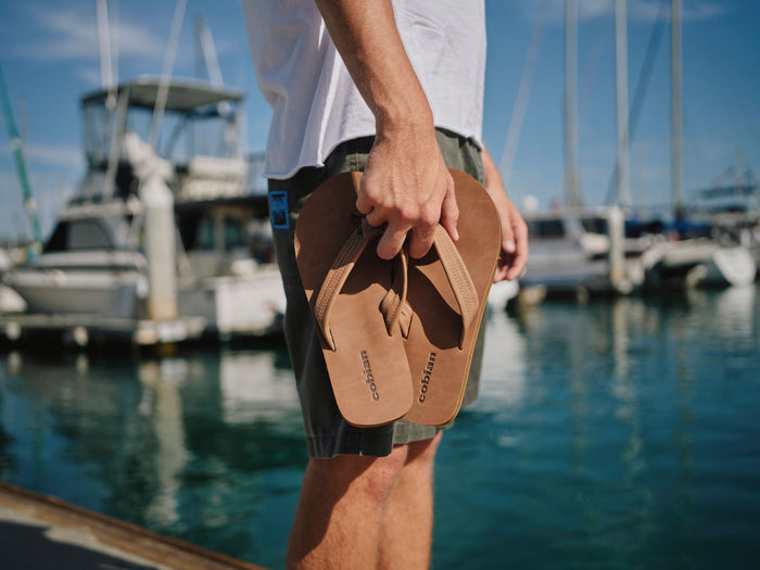 A person standing at a marina holds a pair of tan leather Coast brand flip-flops. The background shows blue water, boats, and a sunny sky. #color_tan