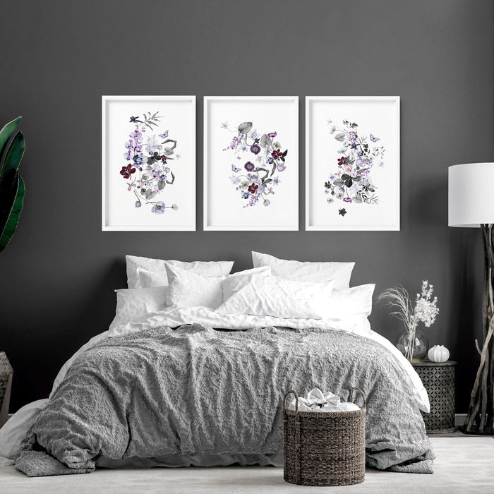 A set of 3 wall art for bedroom featuring delicate purple and burgundy botanical prints in white frames above a grey bed